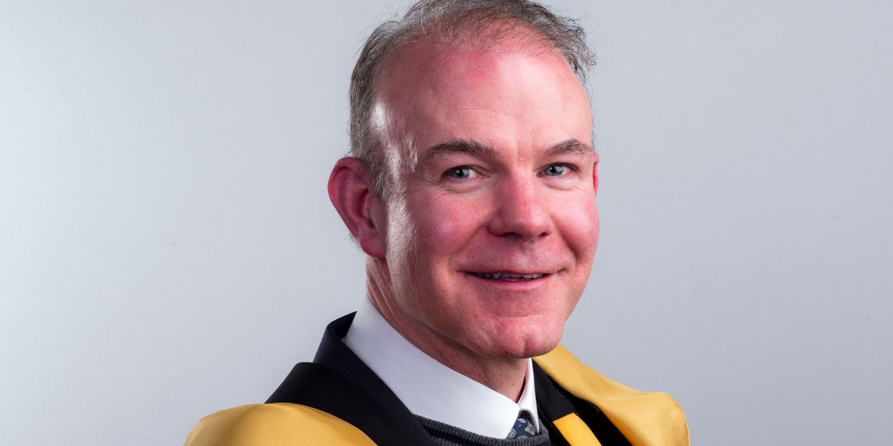 A headshot of a man with grey hair, against a grey background, wearing the yellow and green robes of the Royal Irish Academu